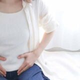 女性が朝、ベットの上でお腹を押さえている。便秘のイメージ。