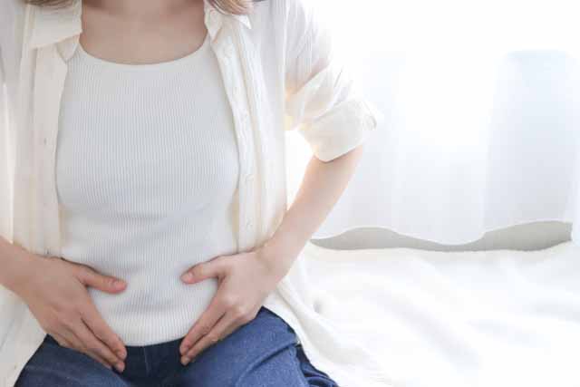 女性が朝、ベットの上でお腹を押さえている。便秘のイメージ。